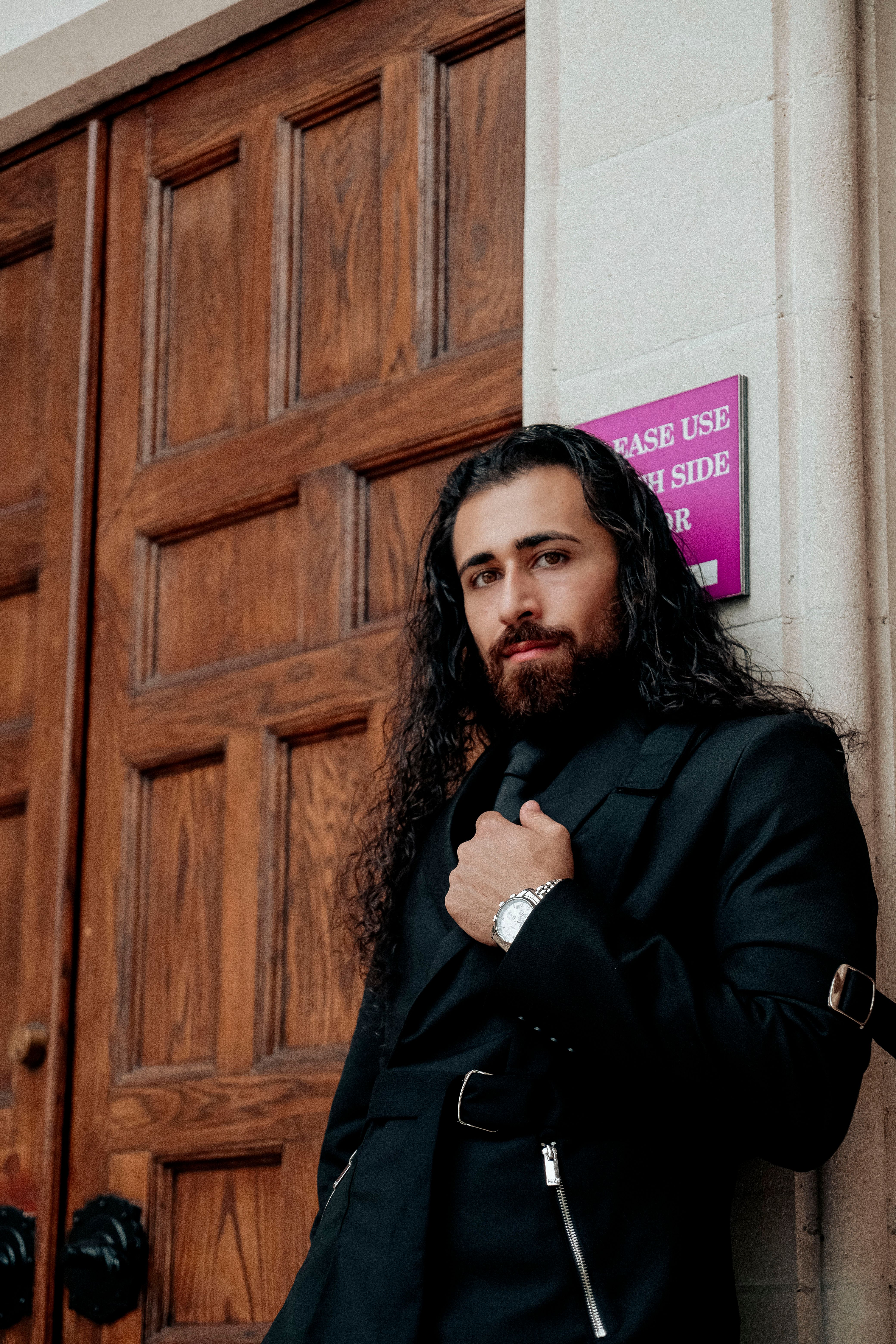 Confident man with long hair leans against a wooden door. The sign adds a vintage touch.