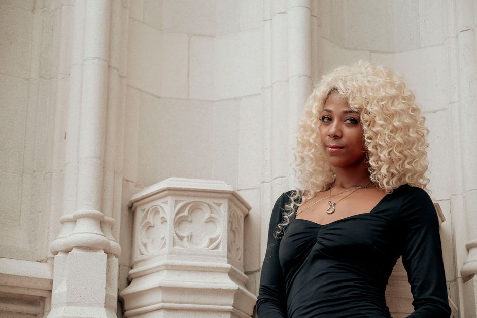 Elegant woman with blonde curls poses in front of detailed stone architecture, showcasing modern style.