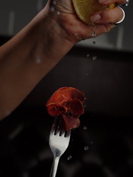 A hand squeezing lemon over chorizo on a fork, with water droplets splashing.