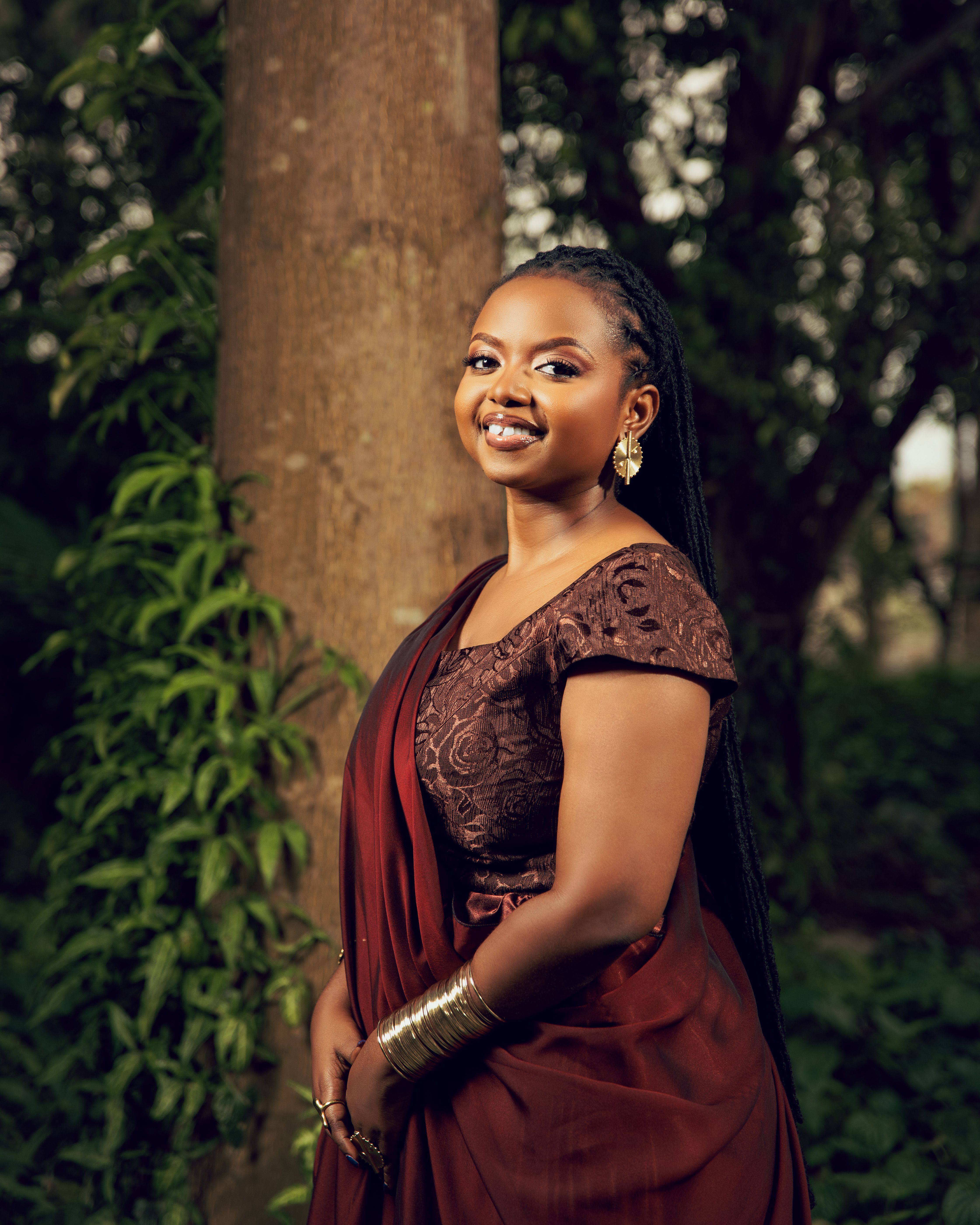 Portrait of a smiling woman in traditional attire, standing in a lush outdoor setting.