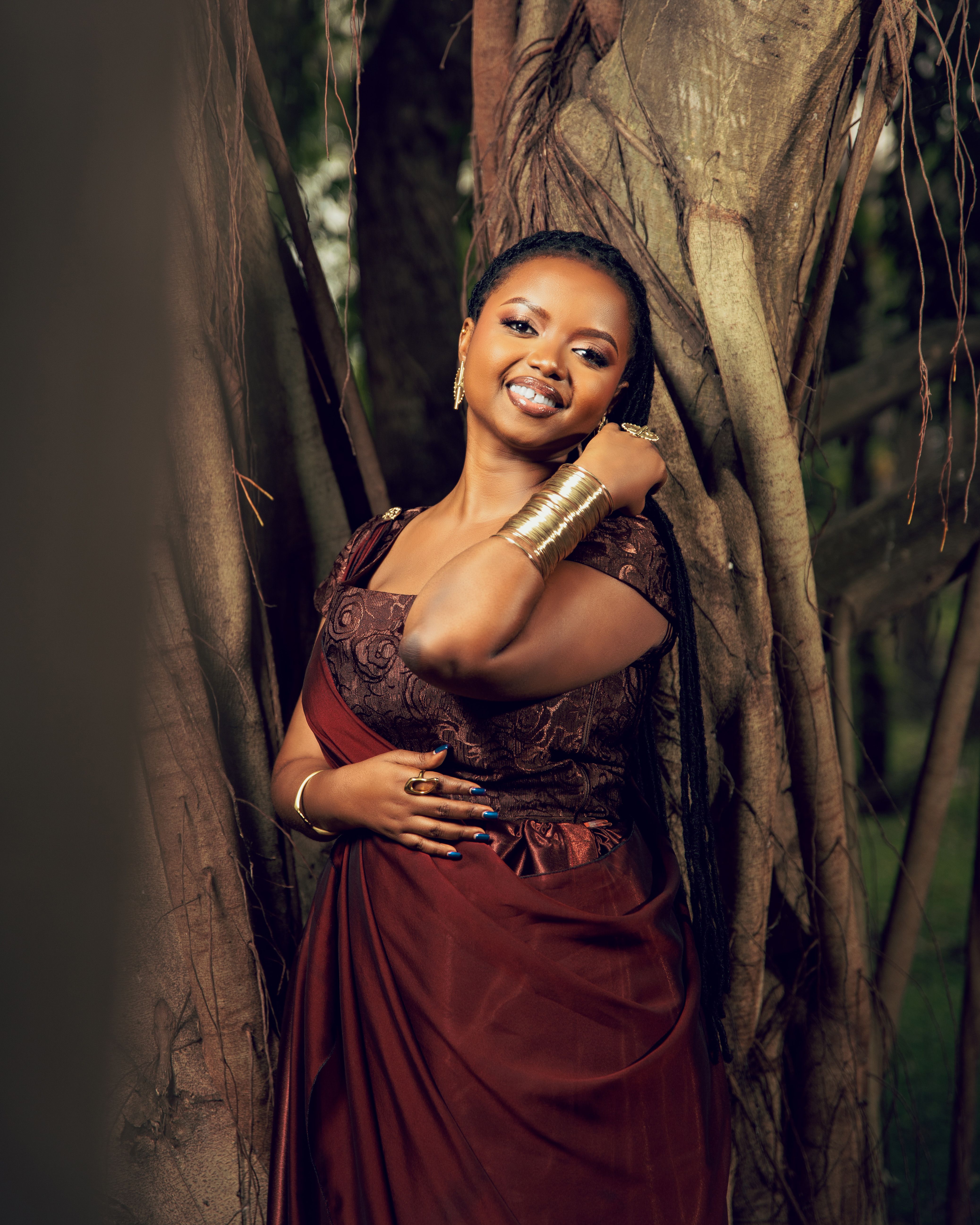 Smiling woman in traditional African attire, standing by a tree, showcasing elegance and cultural beauty.