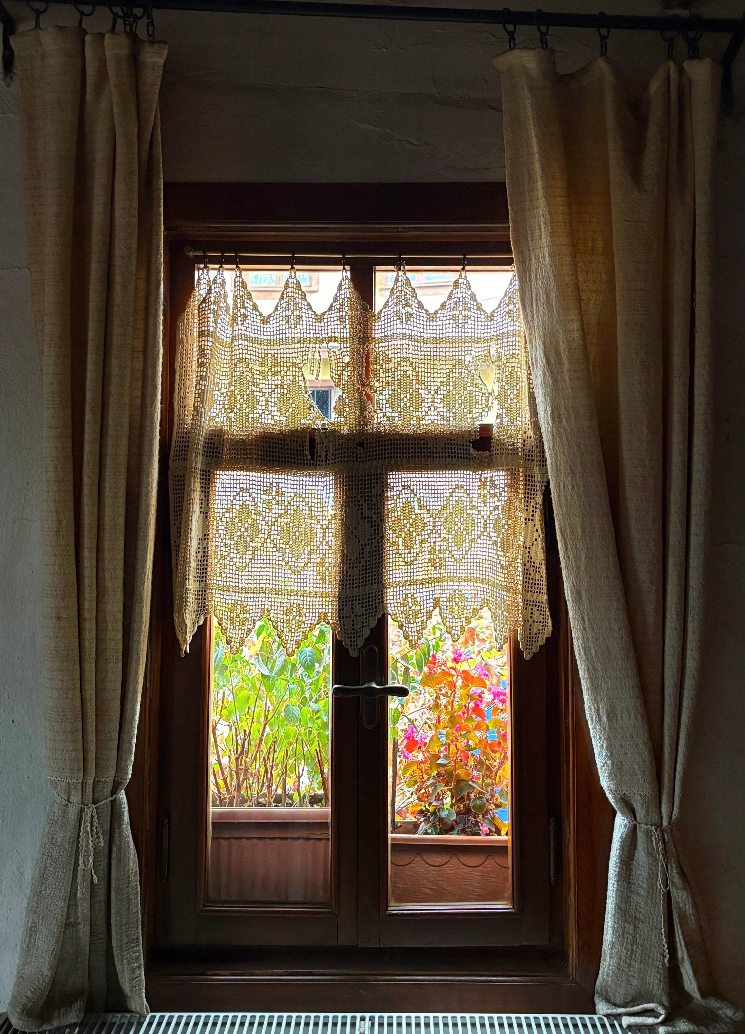 Charming view through a wooden window with lace curtains and colorful flowers outside.