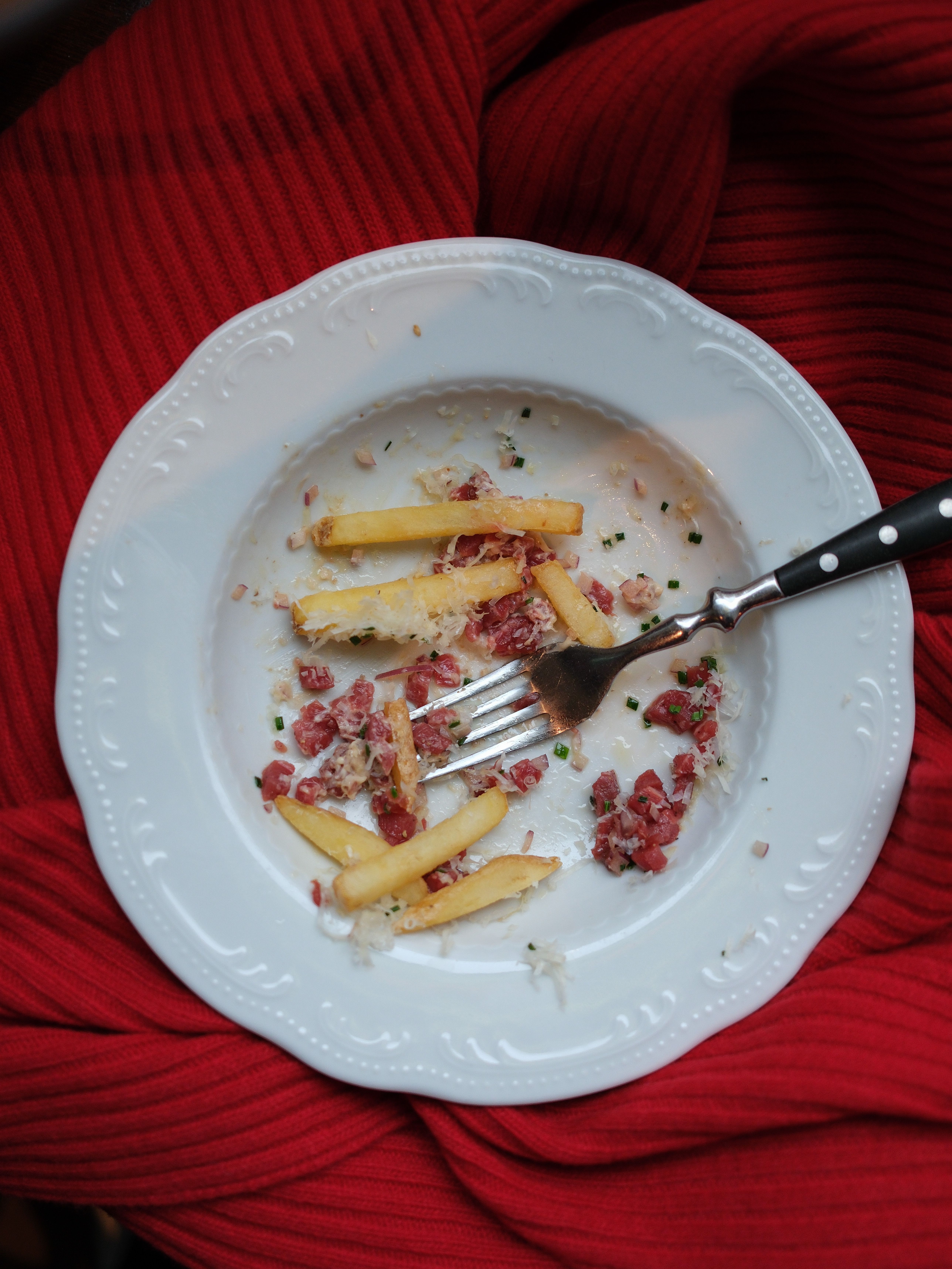 An elegant white plate with leftover French fries and minced meat on a red cloth.