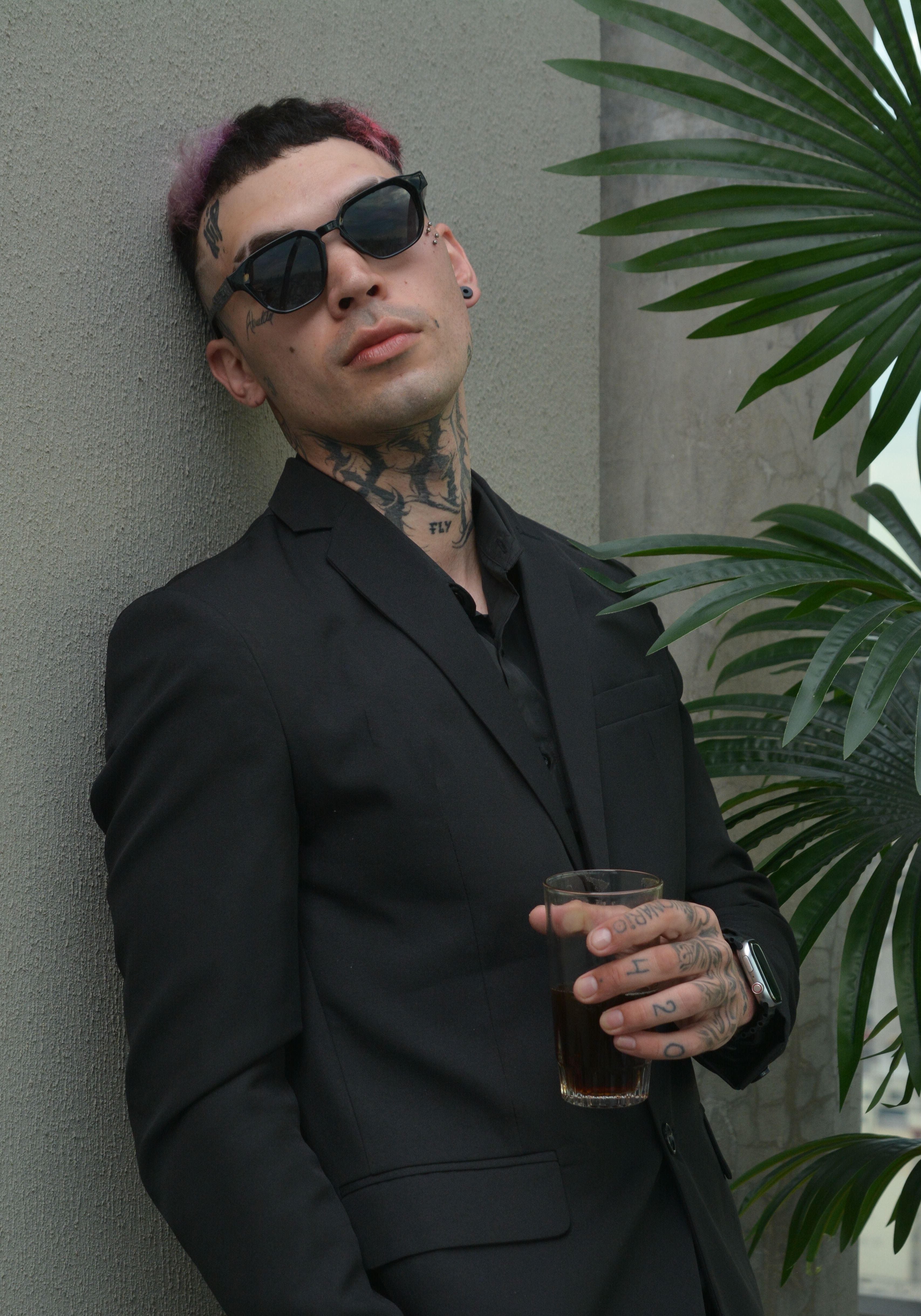 grátis Homem estiloso com tatuagens, terno preto e óculos escuros, segurando uma bebida, exalando confiança e estilo urbano. Foto profissional