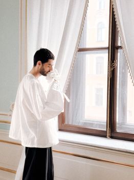 Stylish man in white outfit holding flower by a window in an elegant room.