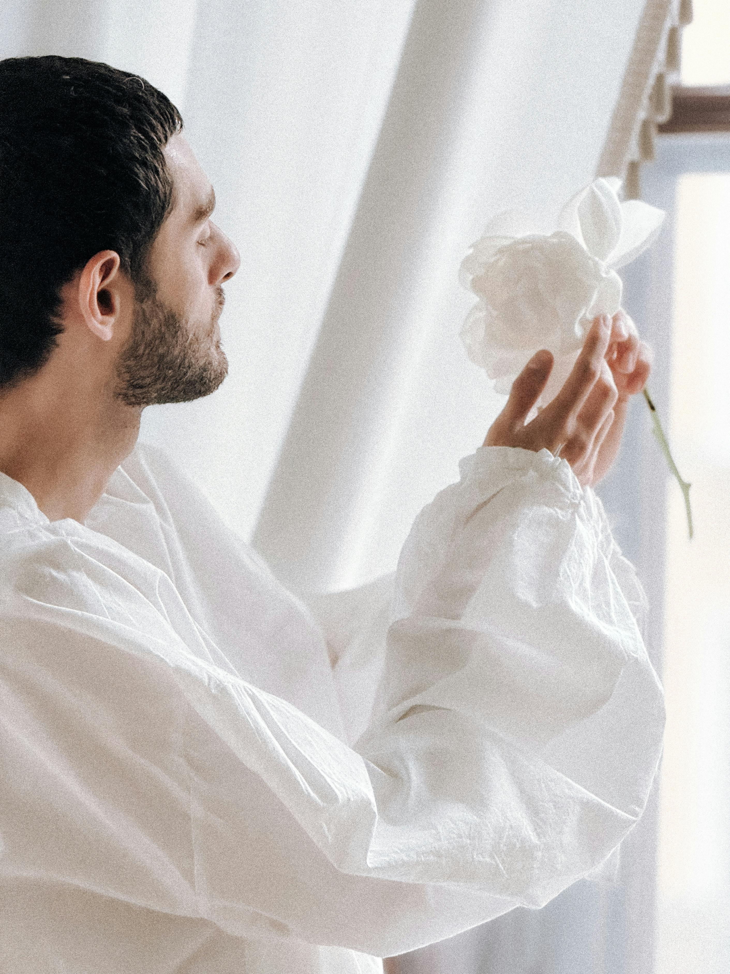 A man admiring a white flower near a bright window indoors.