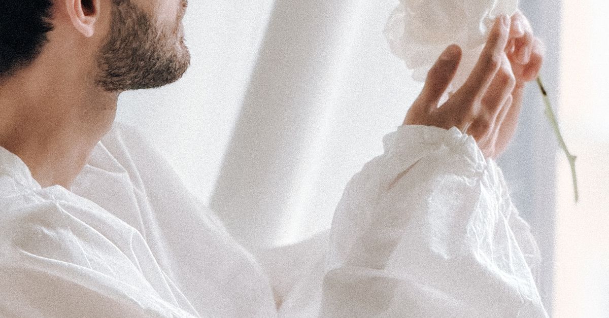 A man admiring a white flower near a bright window indoors.