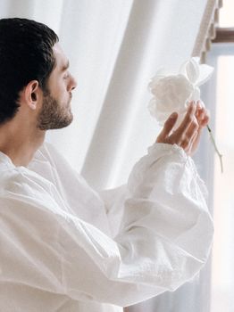 A man admiring a white flower near a bright window indoors.