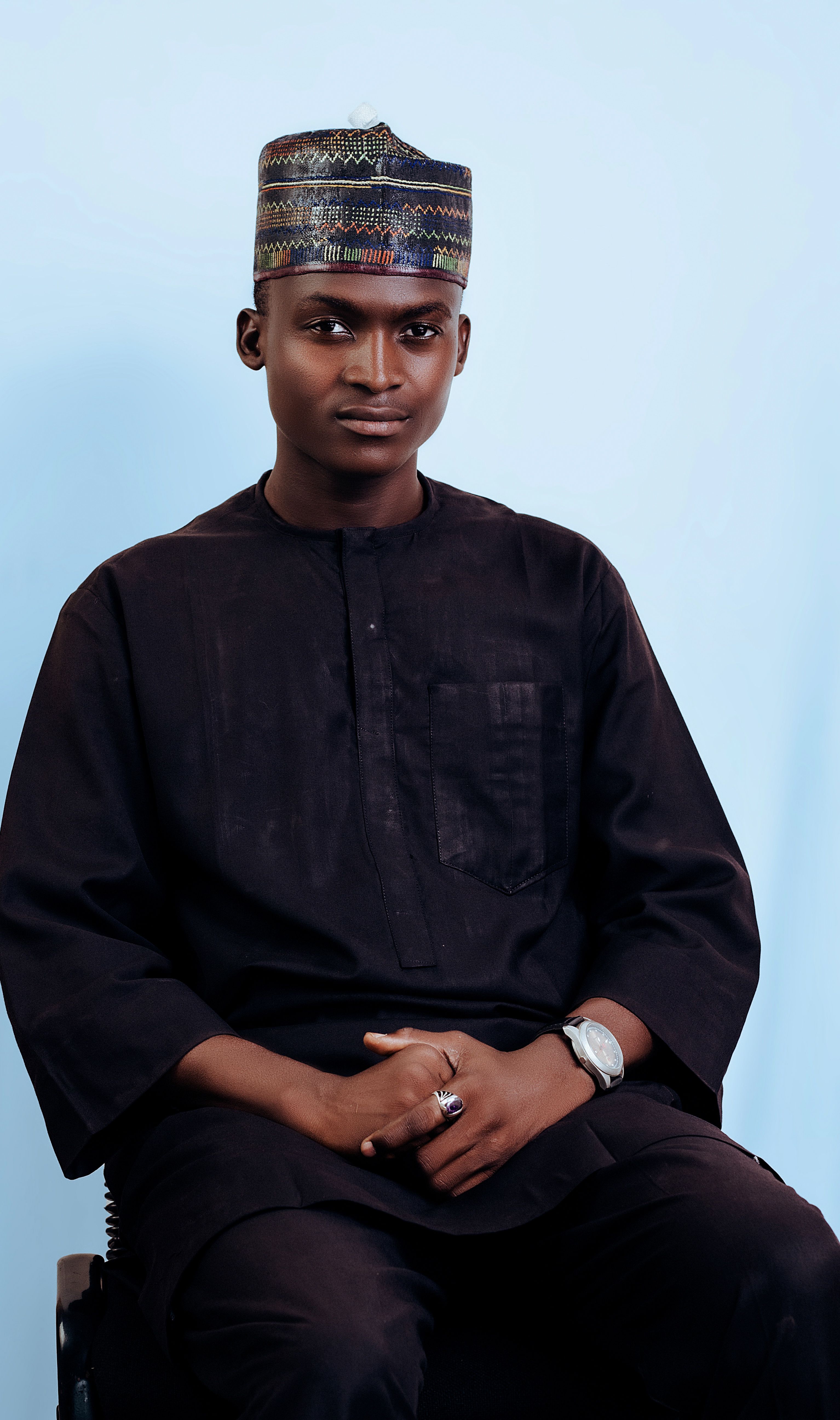 A man in traditional Nigerian attire sits against a blue background, showcasing cultural style.