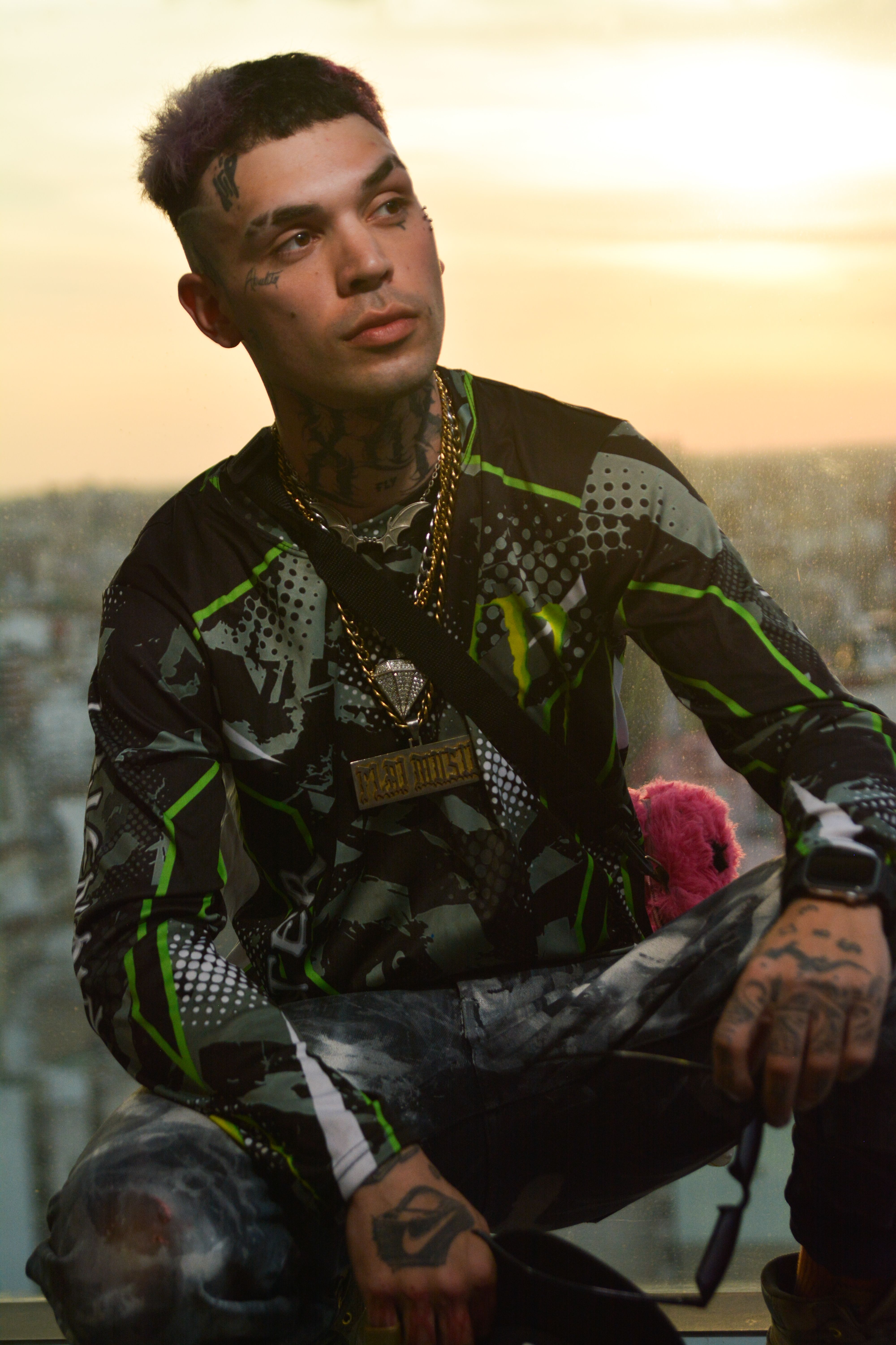 Portrait of a tattooed man in urban wear sitting with a cityscape background at sunset.