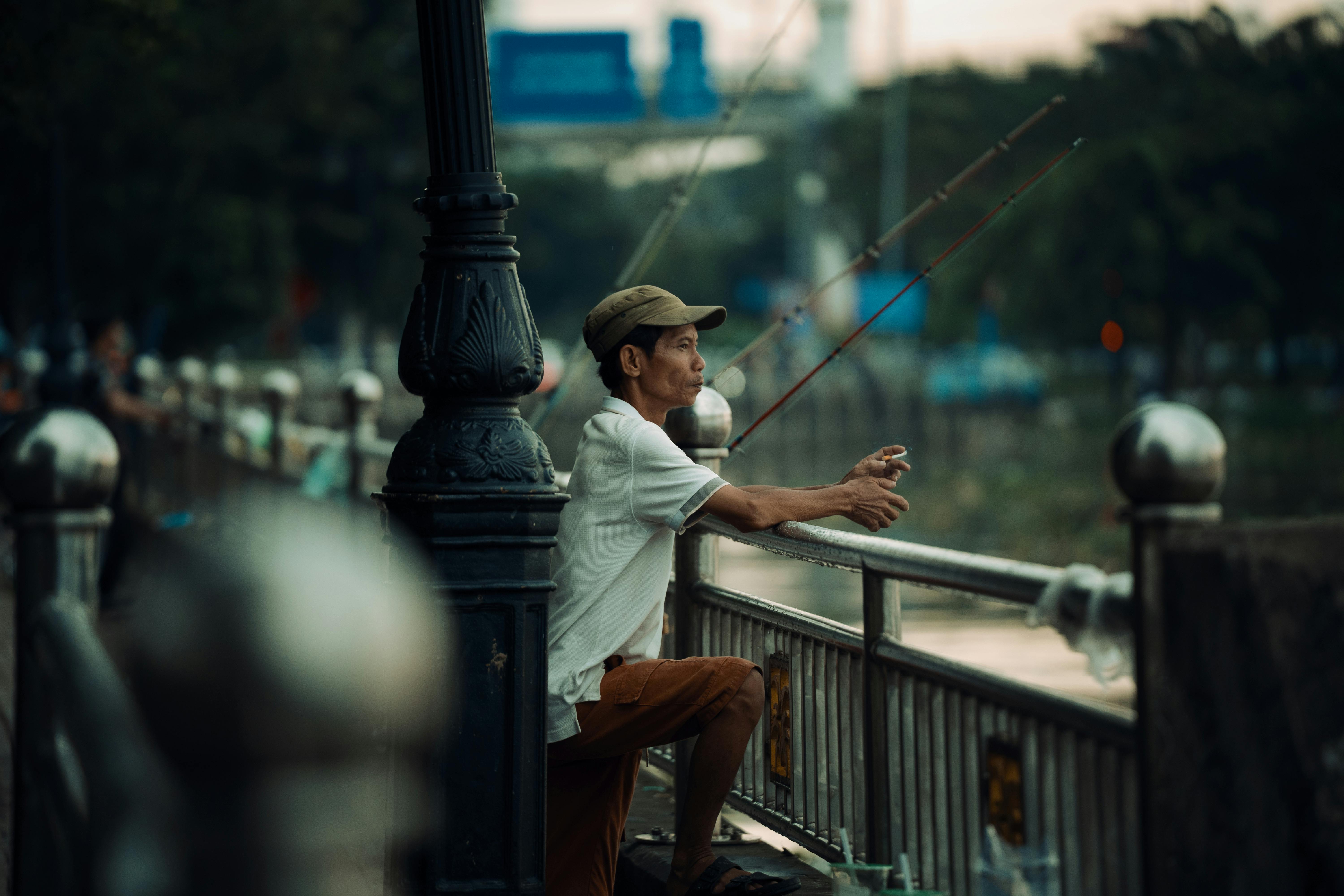 An adult man fishing along riverbank railing at twilight in an urban setting.