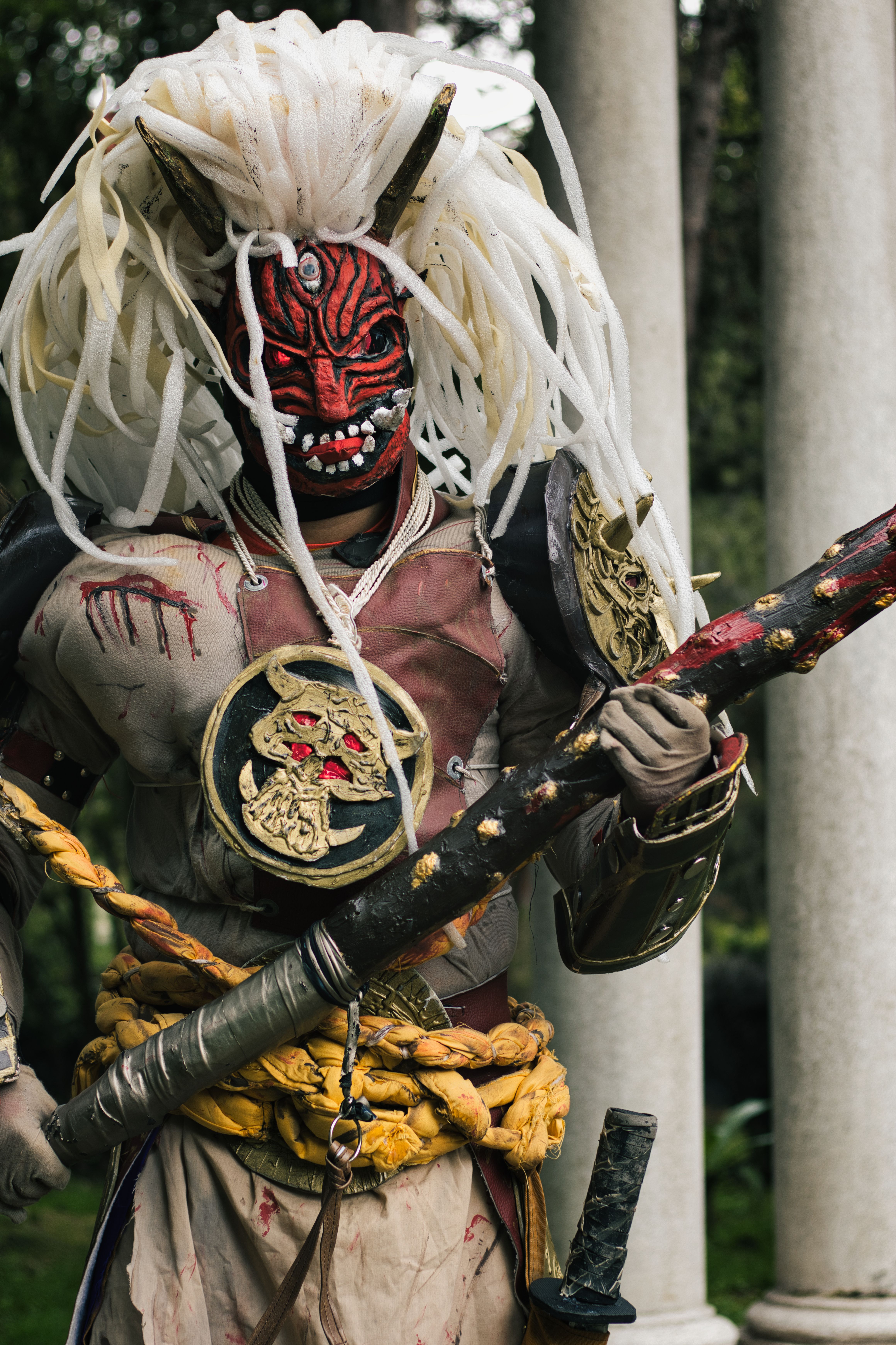 Fierce cosplayer in detailed demon oni armor stands in a lush garden, wielding a menacing weapon.