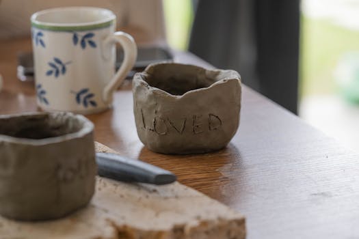 Close-up of clay pottery with 'loved' engraved, next to a mug and tool on a table.