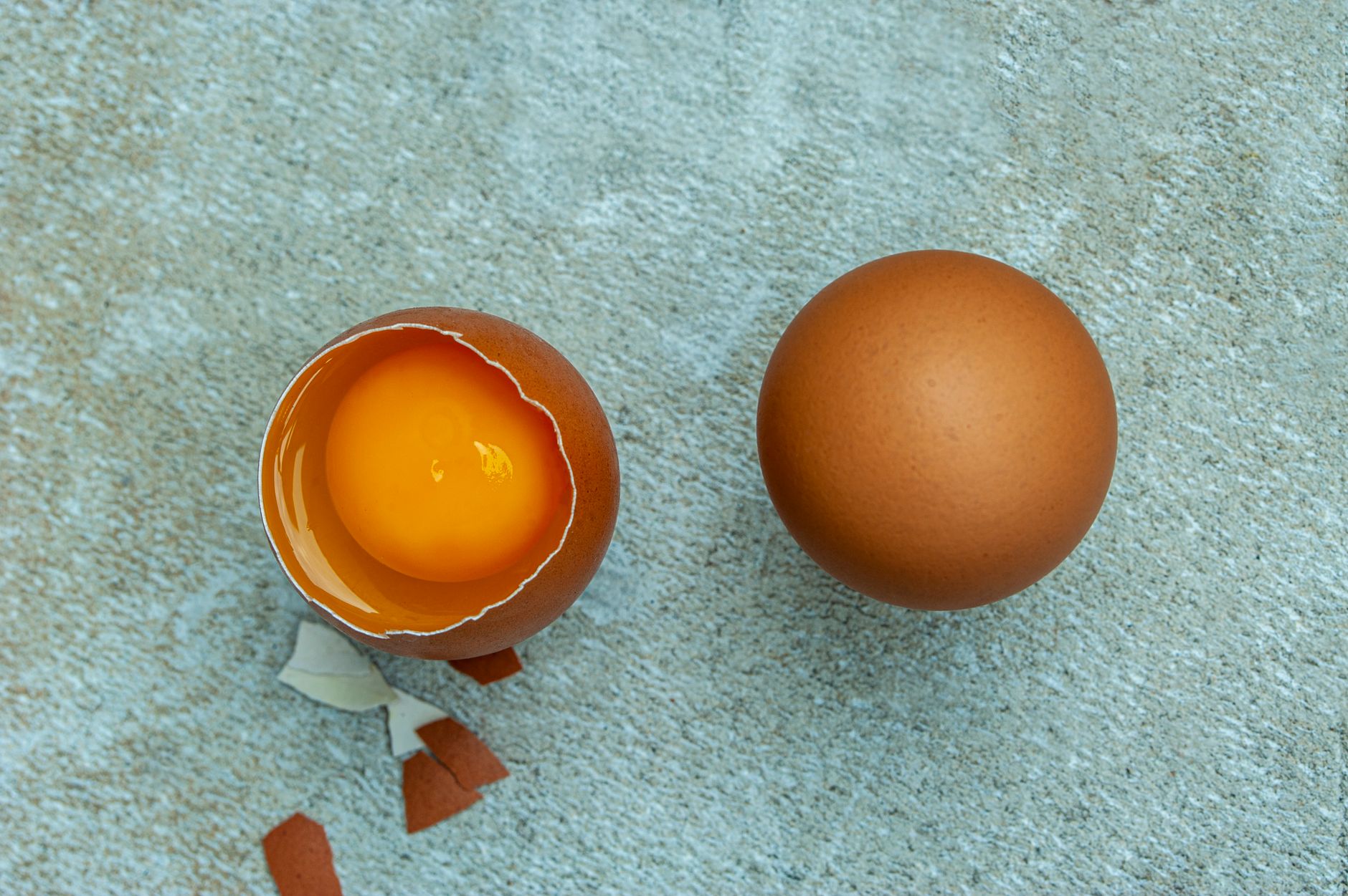 Top view of a cracked brown egg and a whole egg on a textured surface.