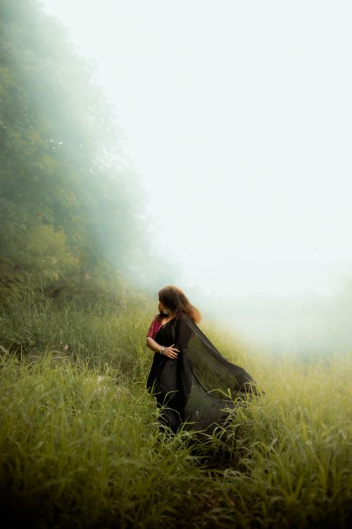 Free A woman in a black dress stands amidst tall grasses enveloped in a mystical, foggy landscape. Stock Photo