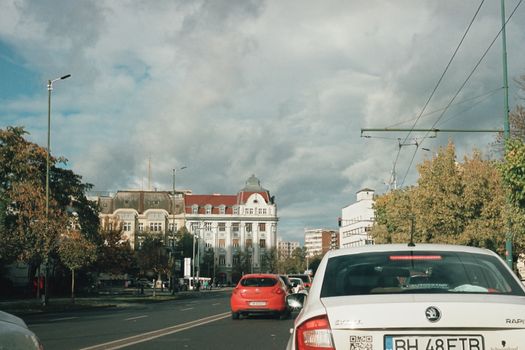 Busy urban street scene featuring cars and a historic building under a cloudy sky.