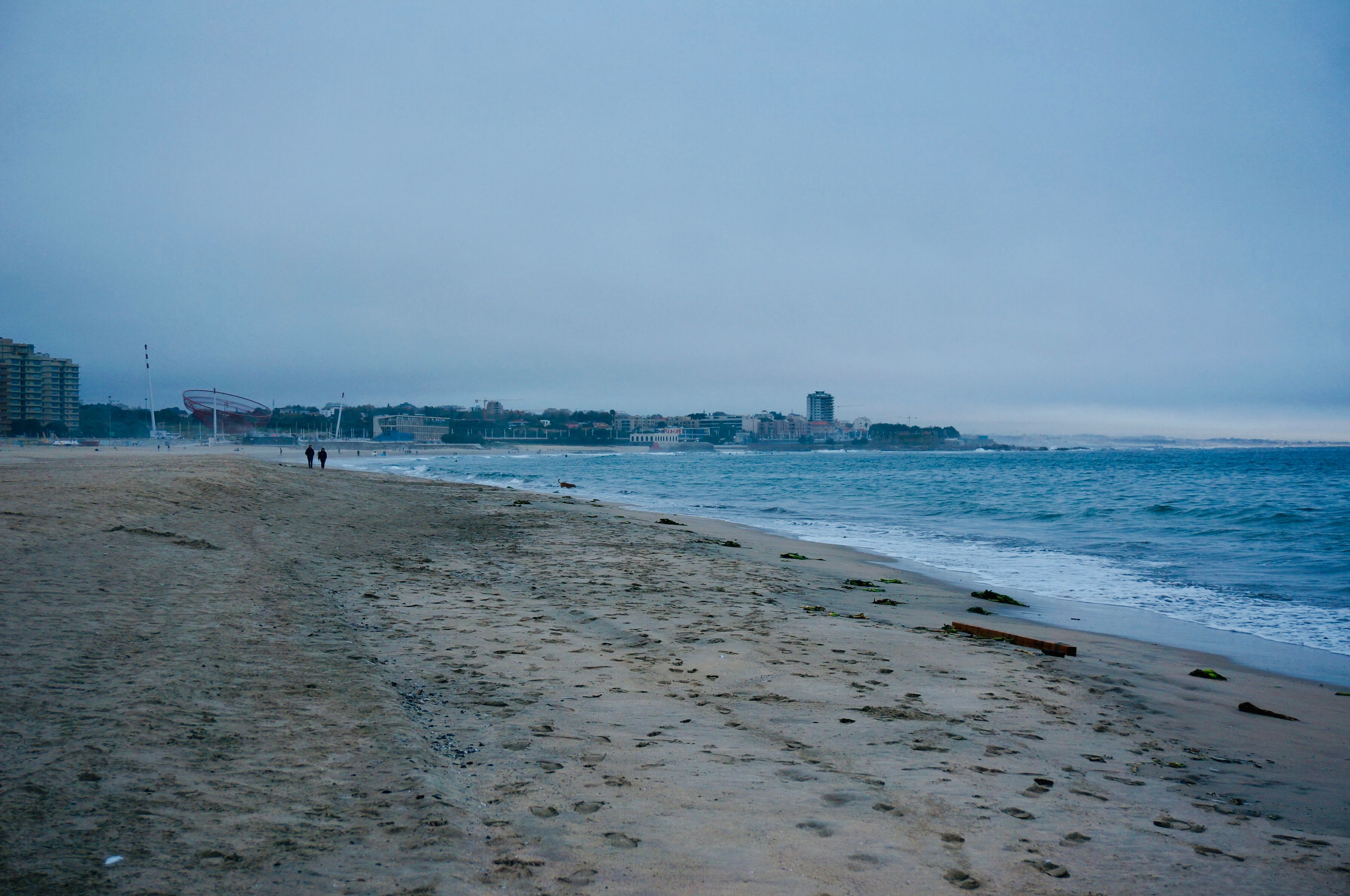 A scenic view of Matosinhos Beach, Portugal, showcasing the sandy shore and distant cityscape under a cloudy sky.