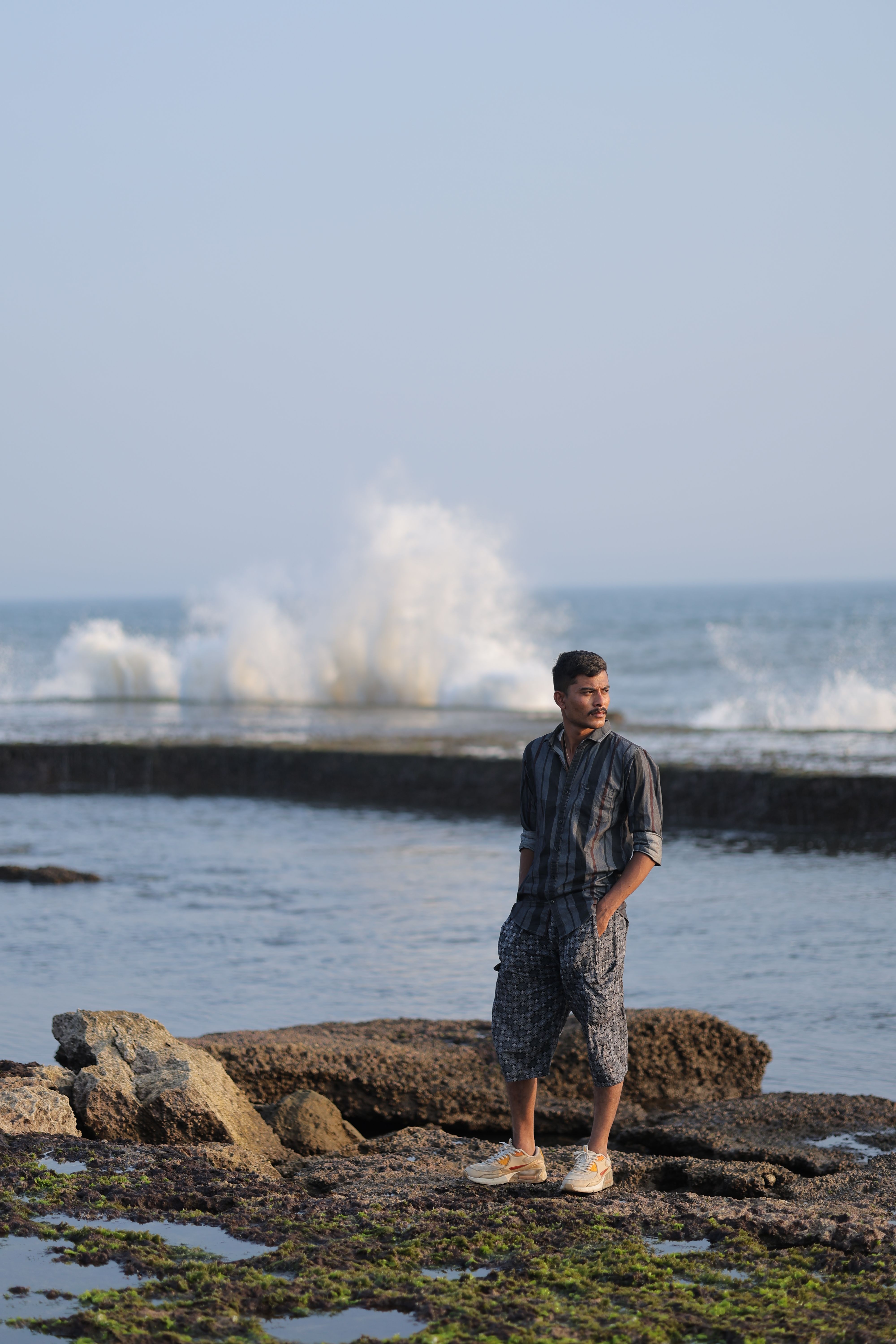 A man in casual attire stands by the ocean as waves crash in the background, creating a serene coastal scene.