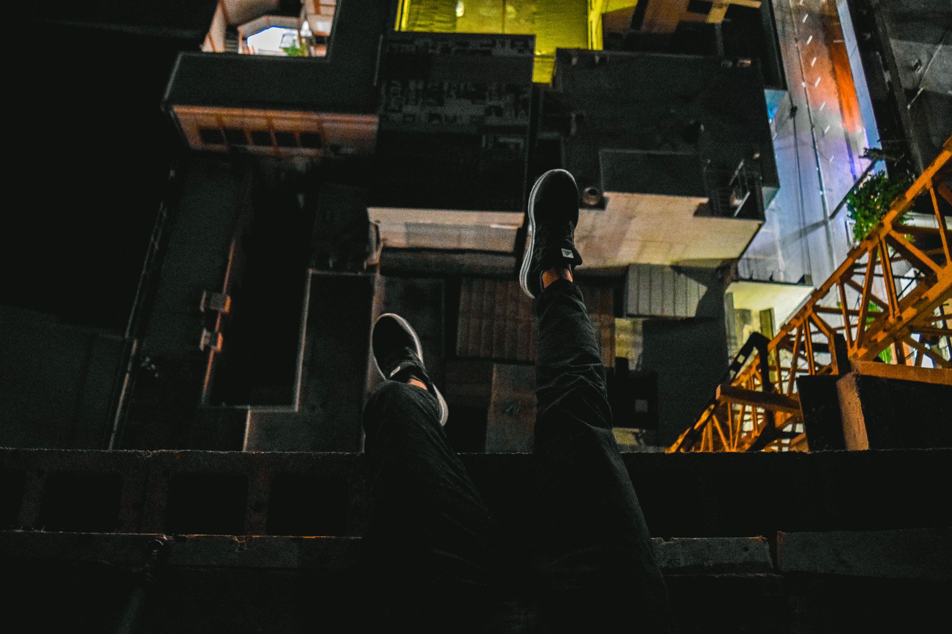 A daring urban rooftop view at night, showcasing a cityscape with legs dangling over the edge. Perfect for adventure or city themes.