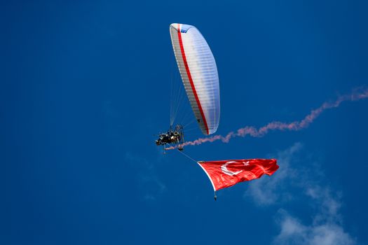 A paraglider flying high in a clear blue sky, adorned with the Turkish flag below.