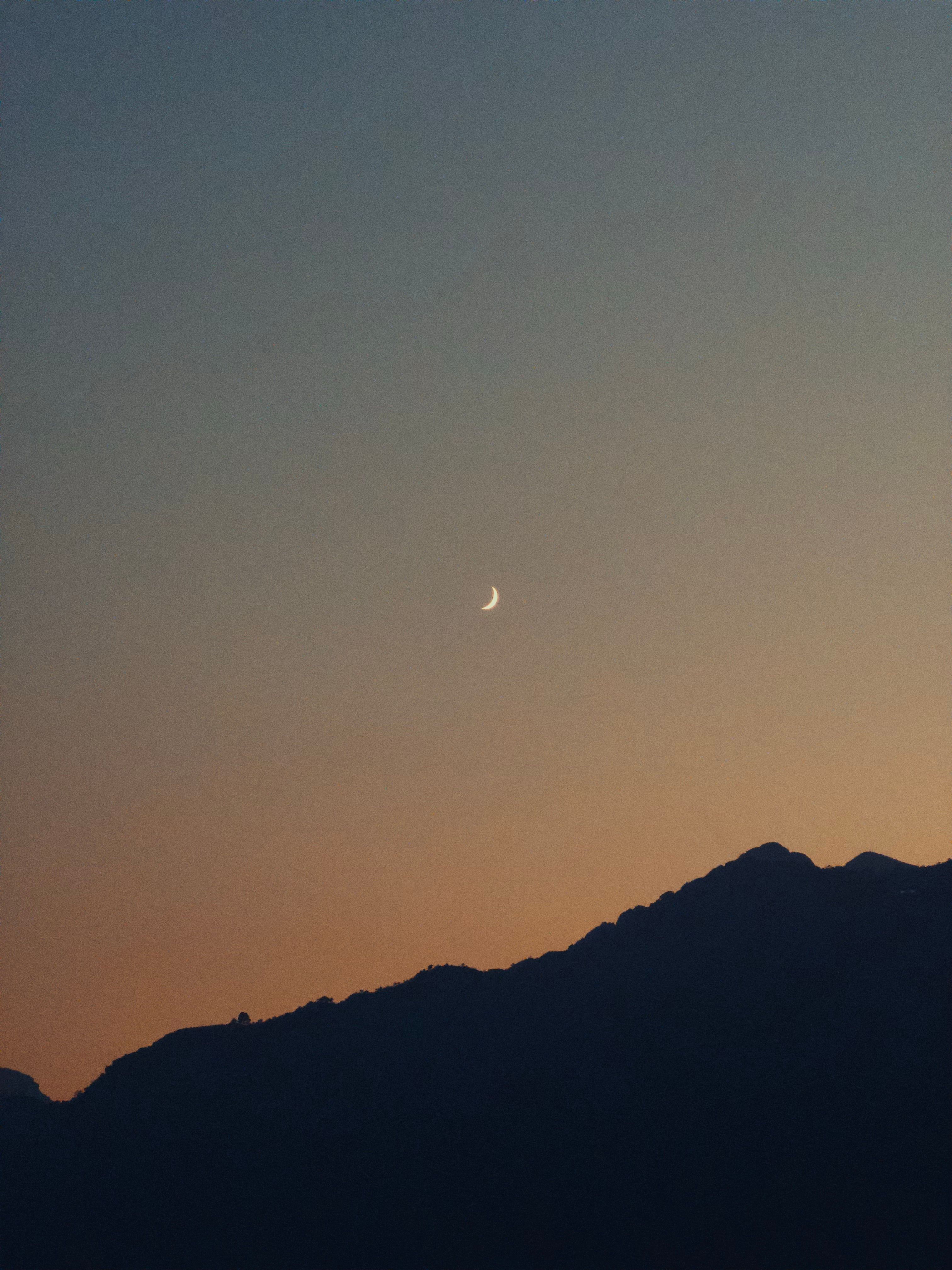 Serene crescent moon above mountain silhouette during dusk twilight.