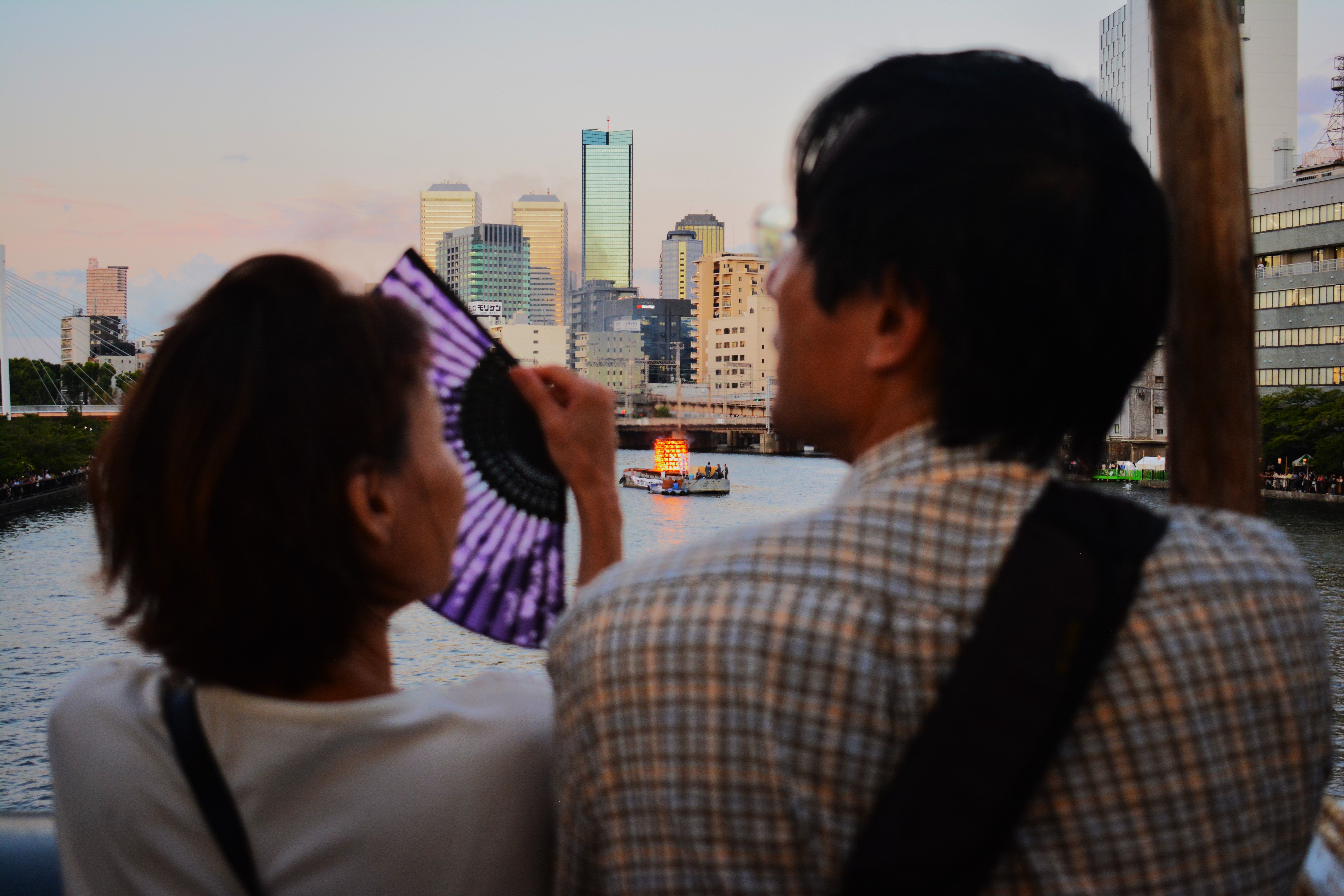A couple enjoys a scenic city view by the river during sunset, with a fan in hand.