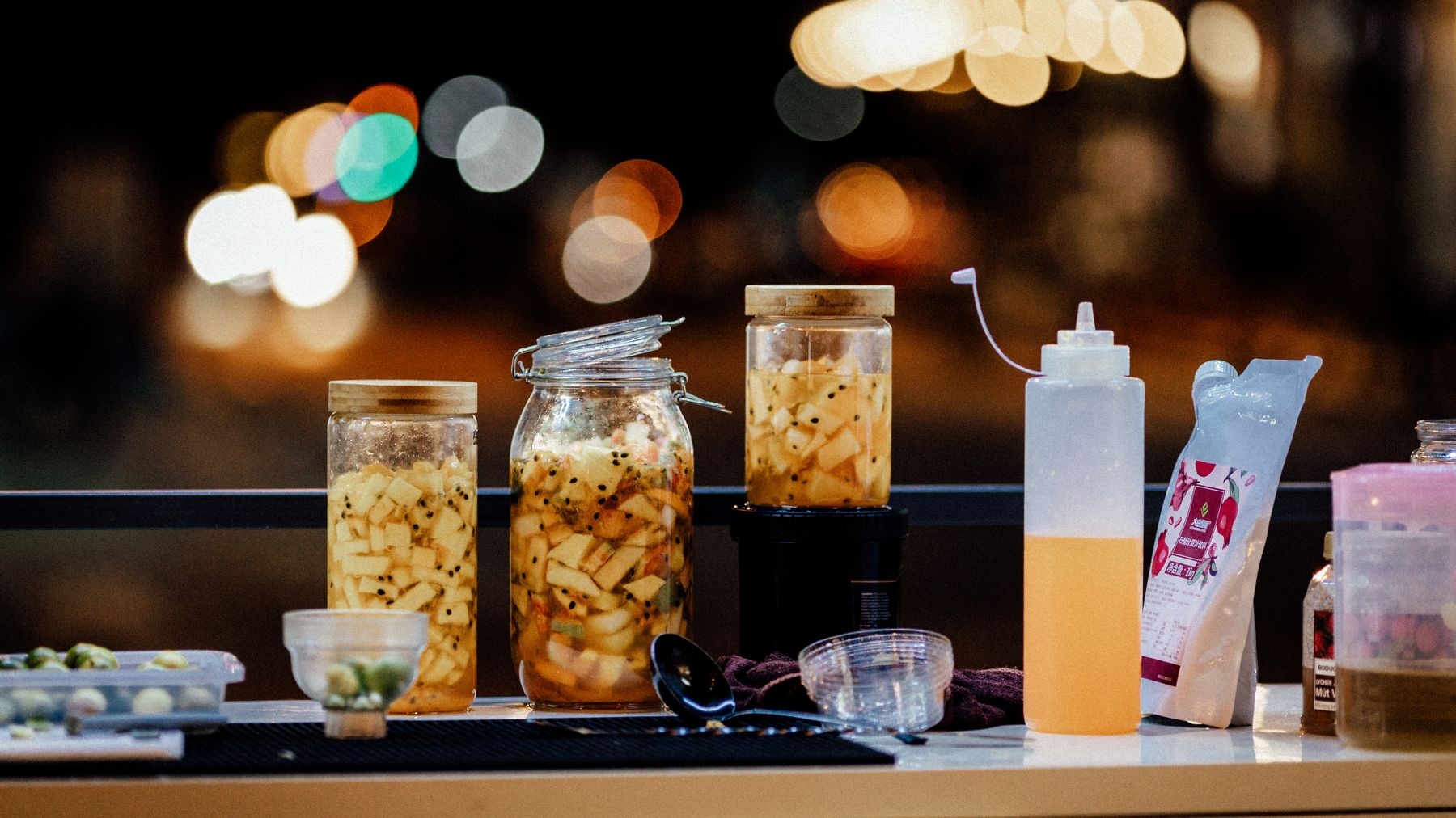 A vibrant display of pickled vegetables in glass jars set at a night market with bokeh lights in the background.