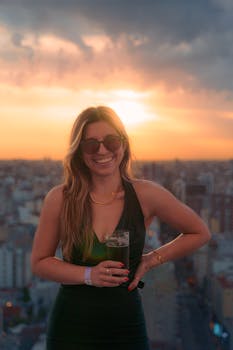 Woman enjoying a sunset with a drink on a rooftop, capturing a vibrant and joyful moment.