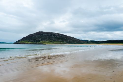 Free Capture of a sandy beach with gentle waves and a lush green hill in the background under an overcast sky. Stock Photo