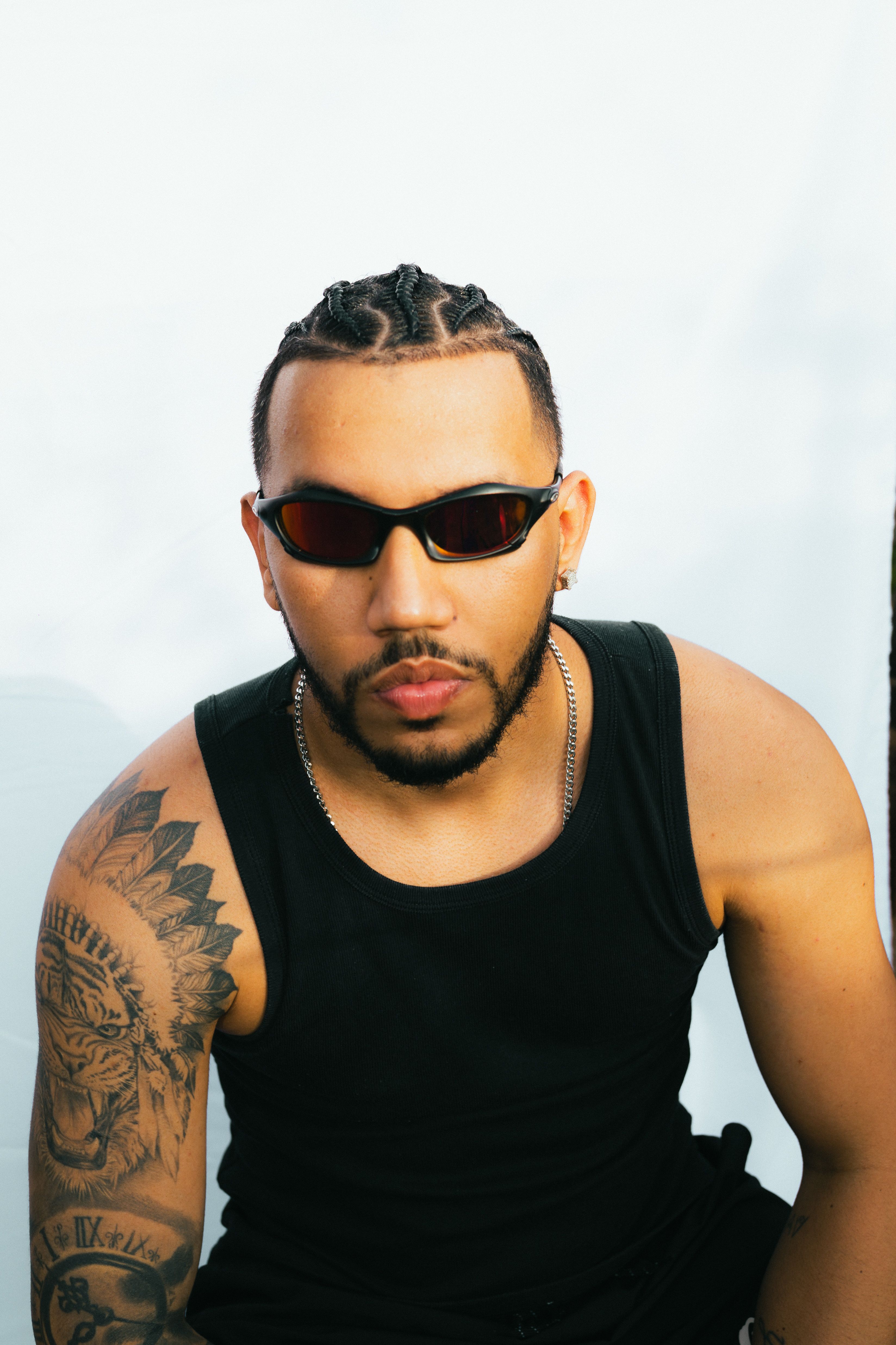 Tattooed man with sunglasses and braided hair posing against a white background.