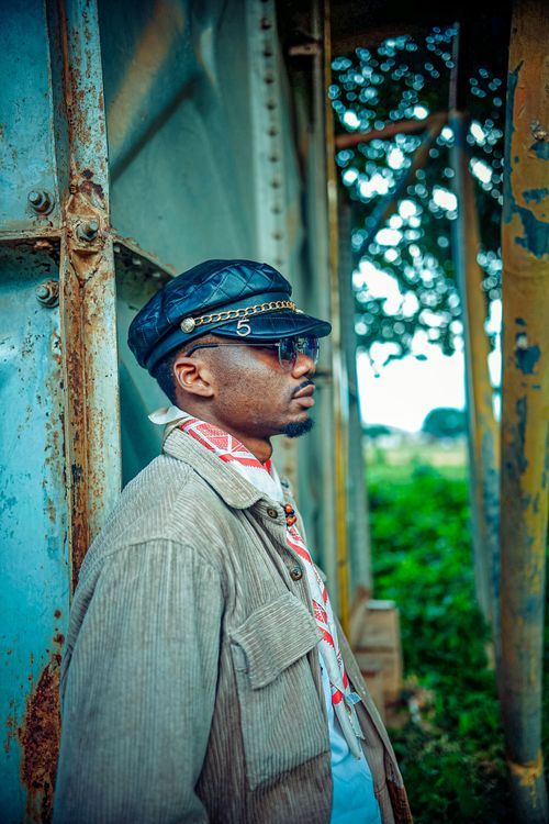 Free Man wearing a cap and sunglasses poses against a metal structure with greenery in the background. Stock Photo