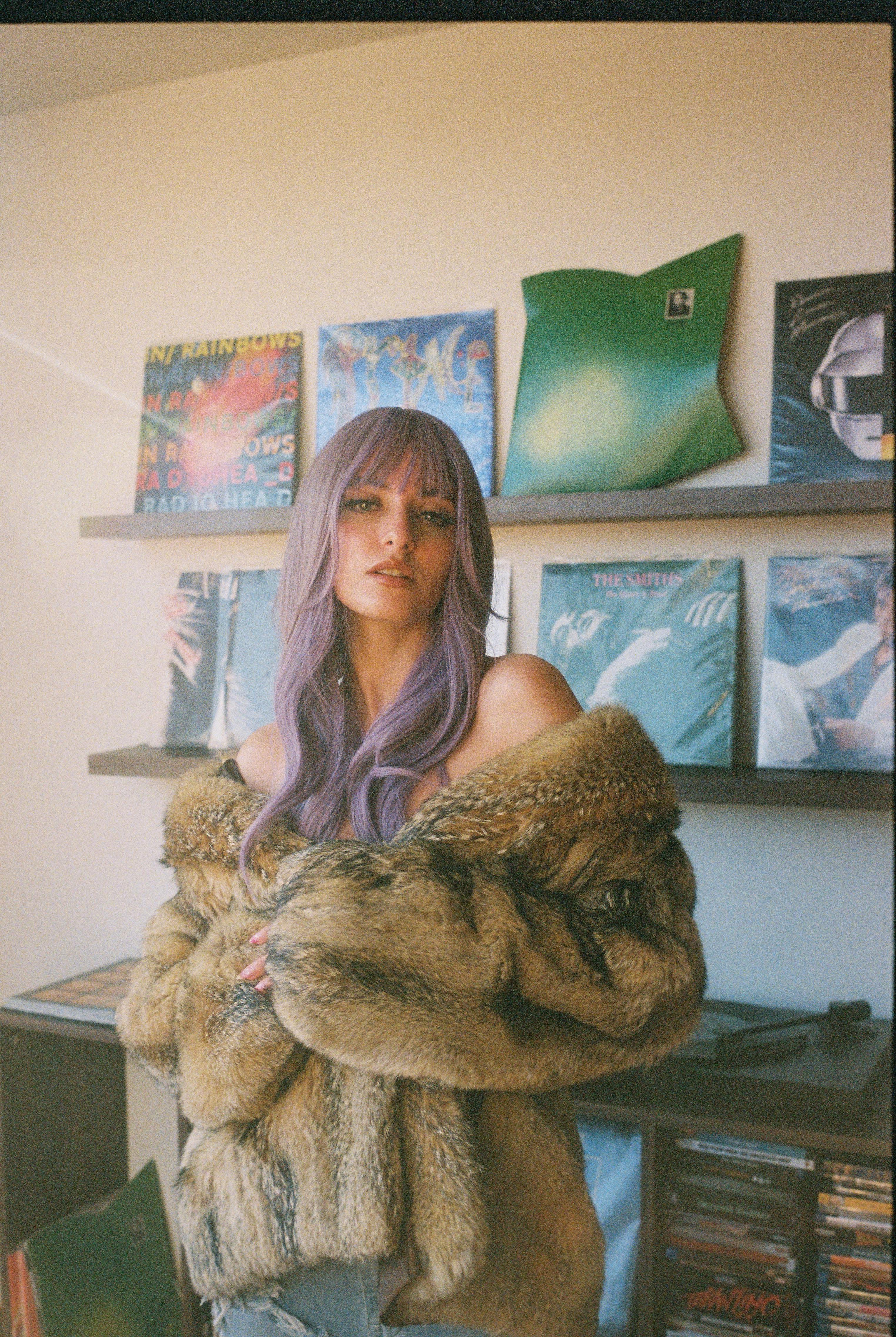 Stylish woman with purple hair in a fur coat standing by vinyl records.