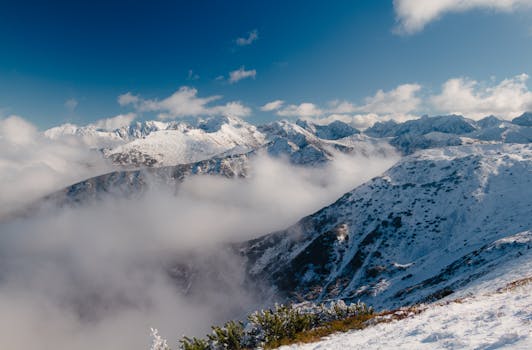 Breathtaking view of snow-covered mountain peaks partially obscured by clouds, under a clear blue sky.
