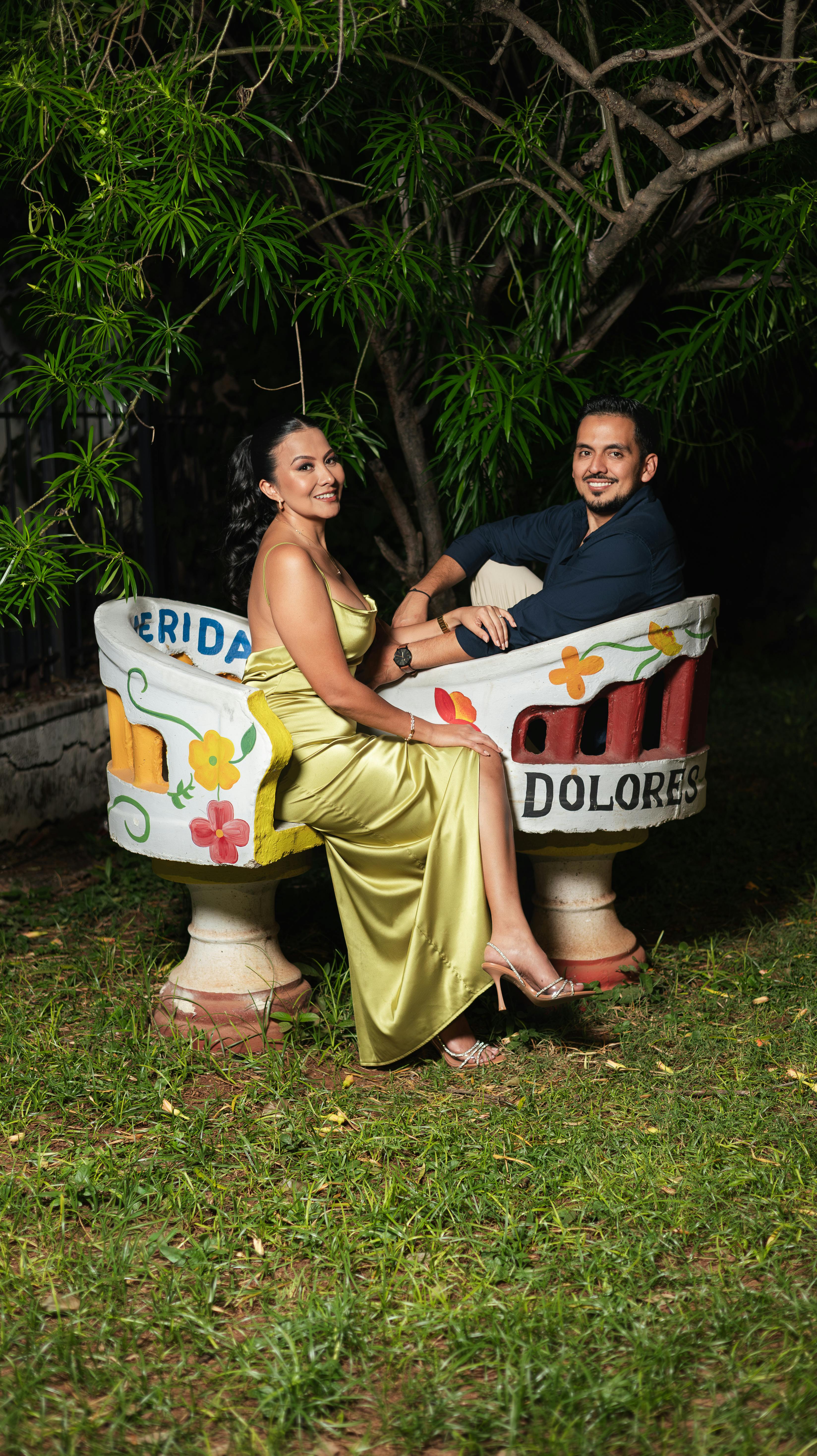 A couple elegantly poses on a decorative bench under lush greenery at night.