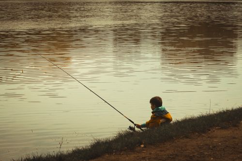 Free A young boy in a yellow jacket fishing by a serene lake in Hungary during a calm day. Stock Photo