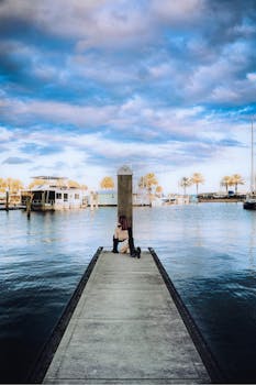 A woman sits on a pier overlooking a tranquil harbor with palm trees and boats, under a dramatic sky.