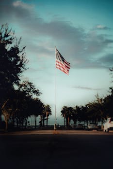 A lone American flag flutters against a vibrant sunset sky, surrounded by trees and a serene ambiance.