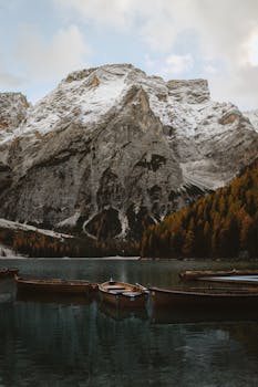 Wooden boats on tranquil Lago di Braies with snow-kissed Dolomites and autumn foliage.