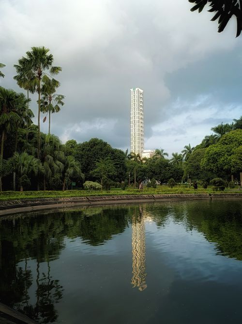 Free A tall skyscraper is reflected in a tranquil park pond surrounded by lush greenery under a cloudy sky. Stock Photo