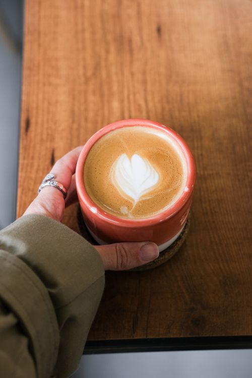 Free Hand holding a latte with heart-shaped foam art in a red cup on a wooden table. Stock Photo