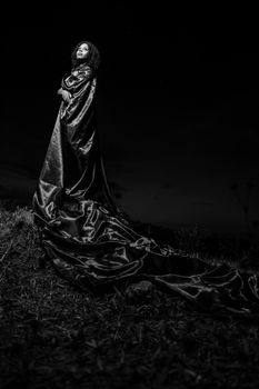 Black and white portrait of a woman in a flowing gown, standing elegantly outdoors at night.