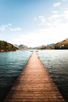 A tranquil wooden dock extends into a serene lake in Austria, surrounded by lush mountains under a blue sky.