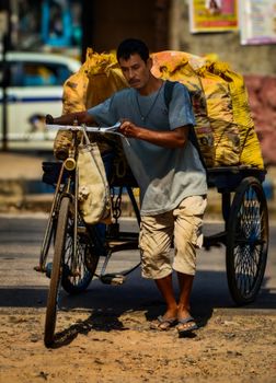 A man resting by his bicycle rickshaw on a busy urban street, loaded with yellow sacks.