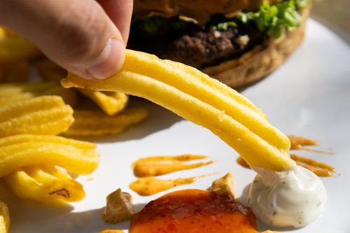 Free Close-up of a crispy fry being dipped in sauces with a burger in the background, showcasing a tasty meal. Stock Photo