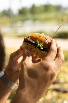 A close-up of two hands holding a burger outdoors in a sunny setting, showcasing fresh ingredients.