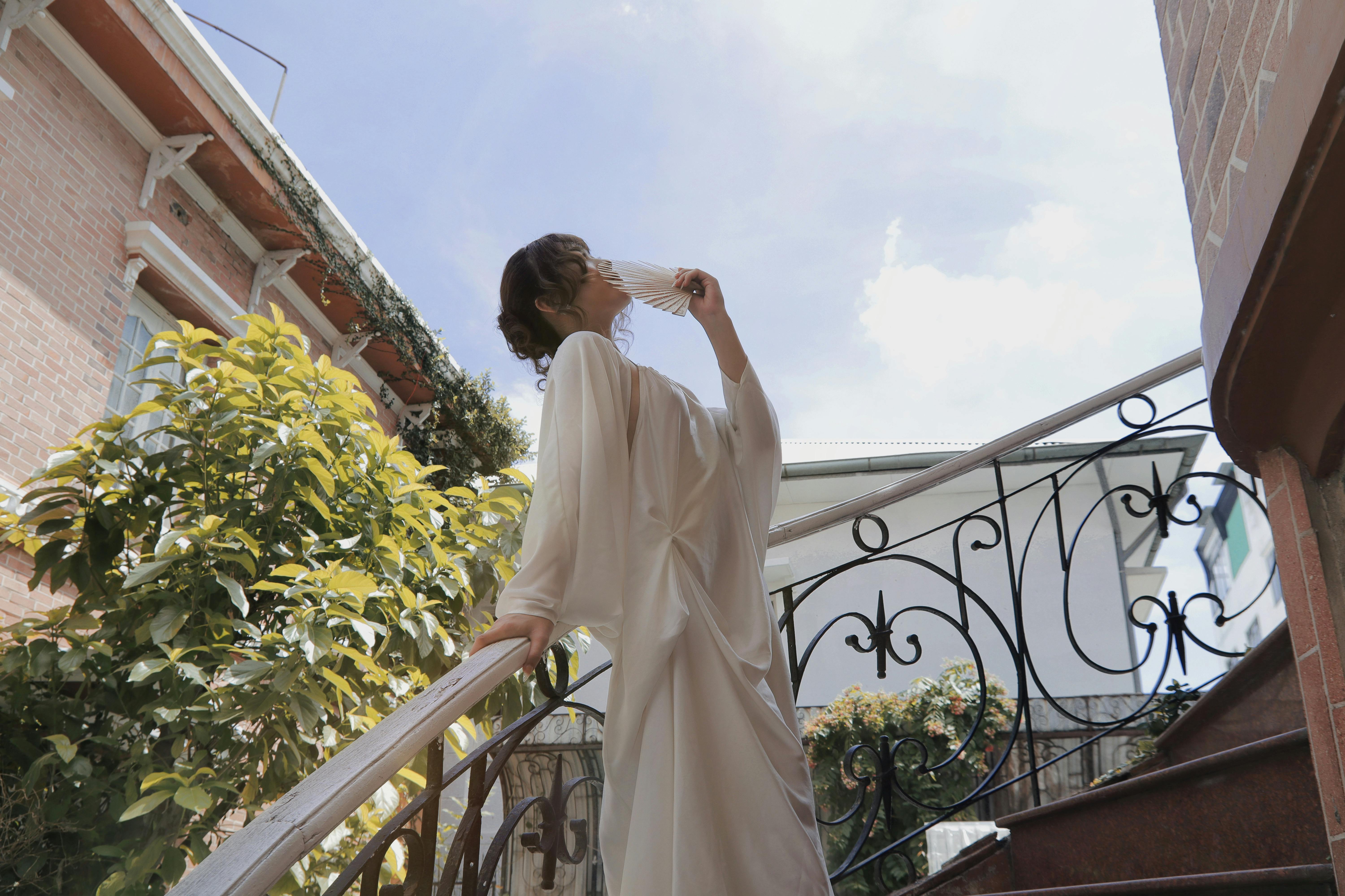 A woman in a flowing dress gracefully poses with a fan on an outdoor staircase under clear blue skies.