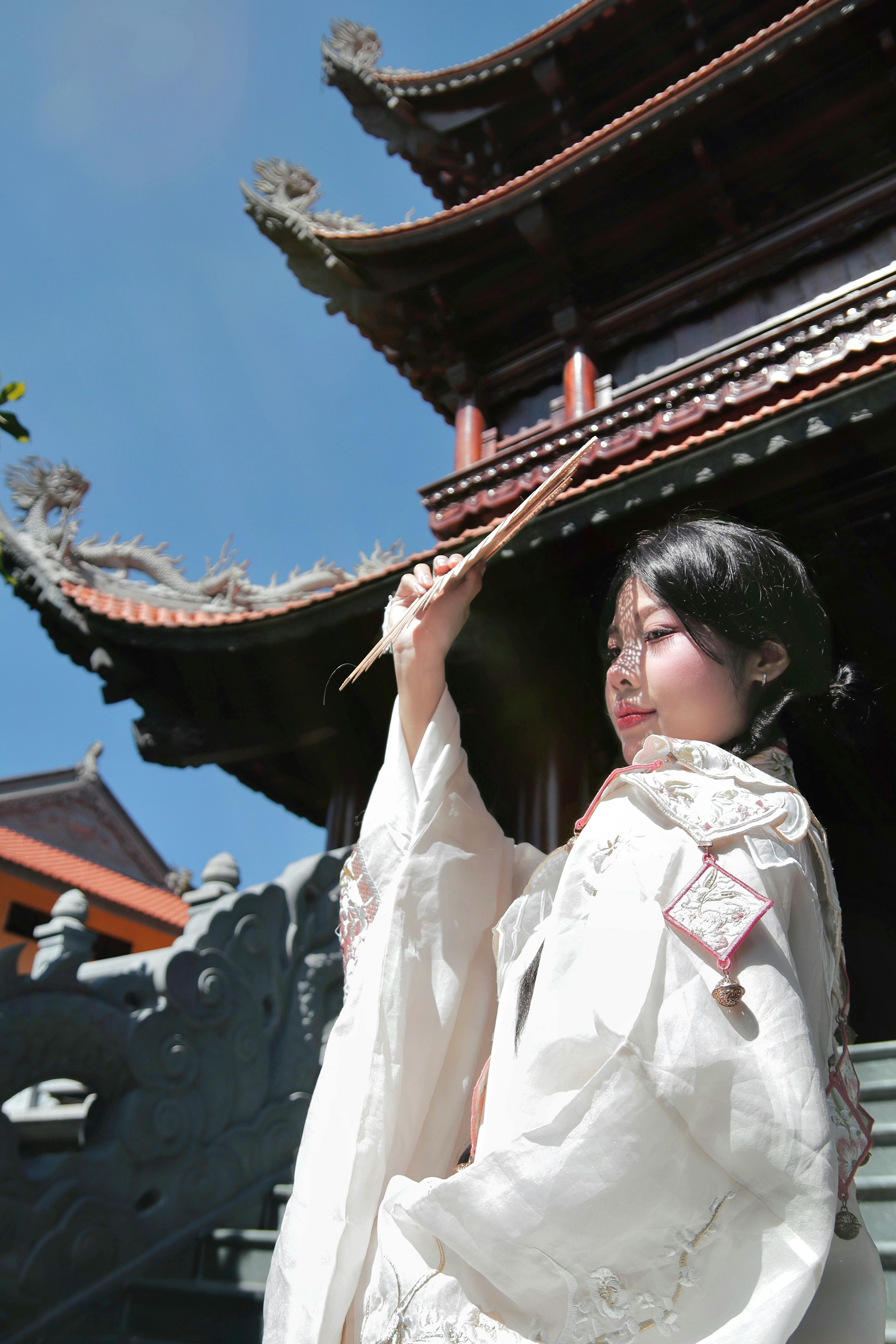 A woman wearing traditional Asian clothing poses outdoors near an ornate temple under the clear sky.