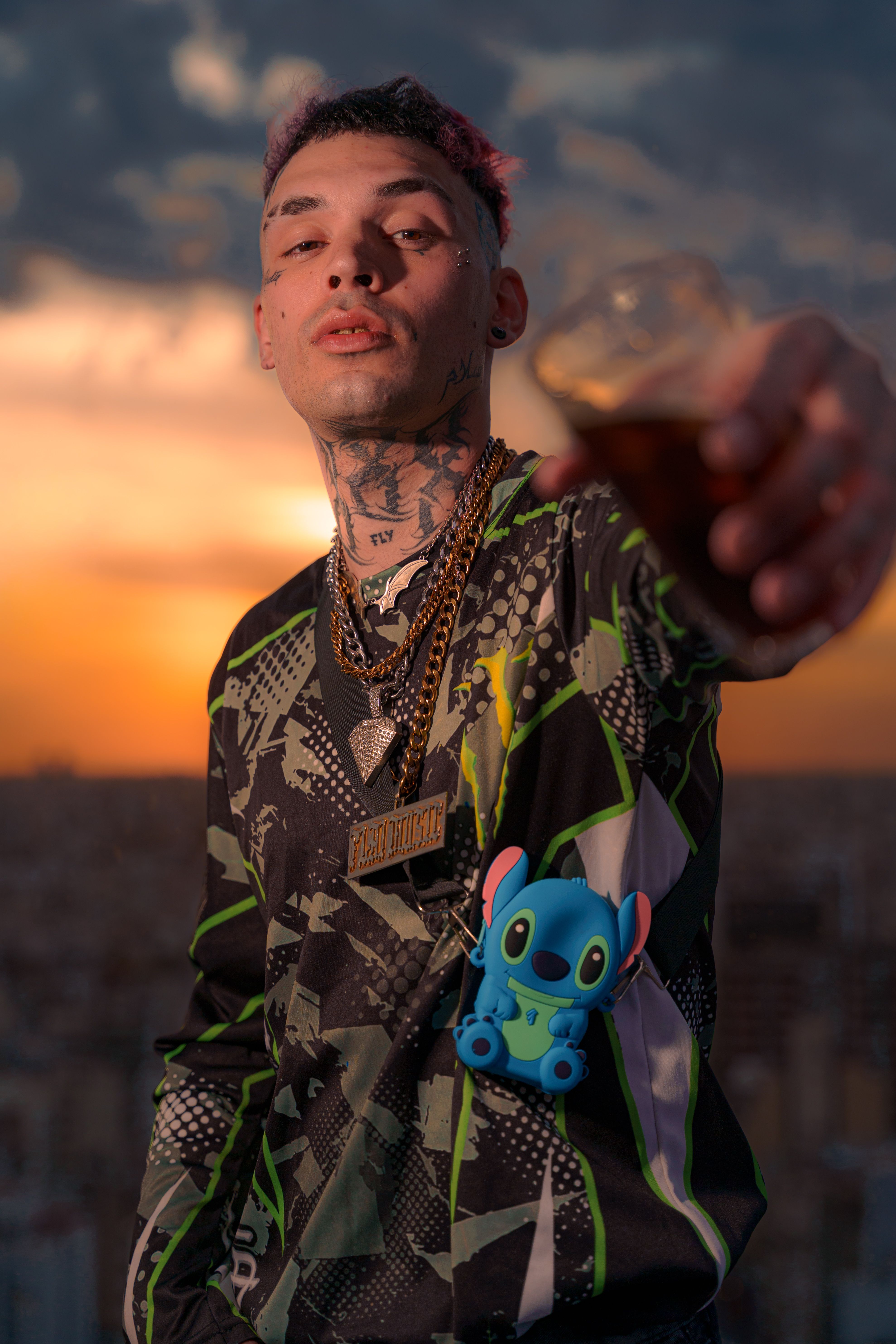 Young adult with tattoos and stylish attire holding a drink at sunset.