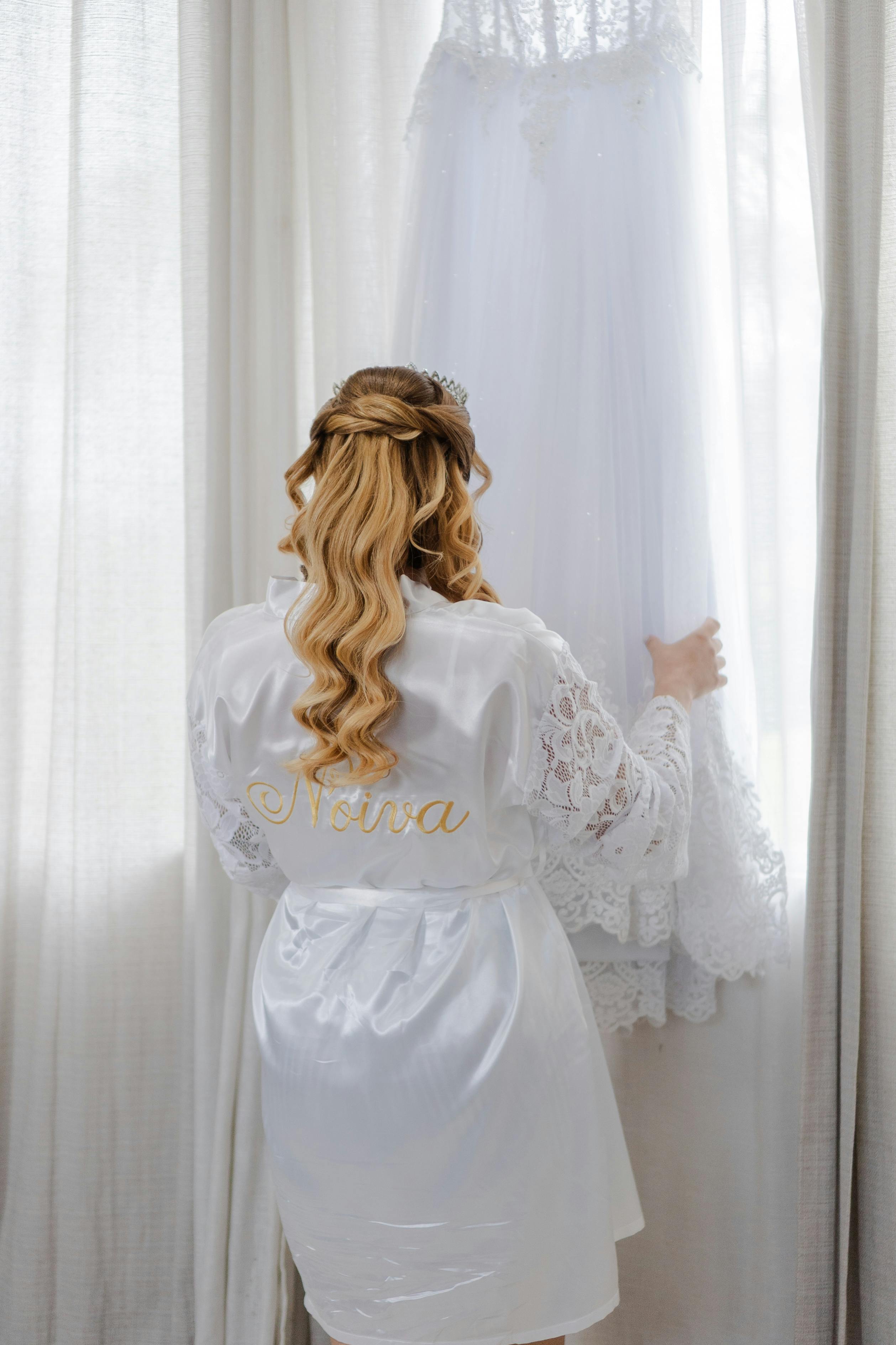 Bride in robe admiring her wedding dress by a sunlit window, creating an elegant morning moment.