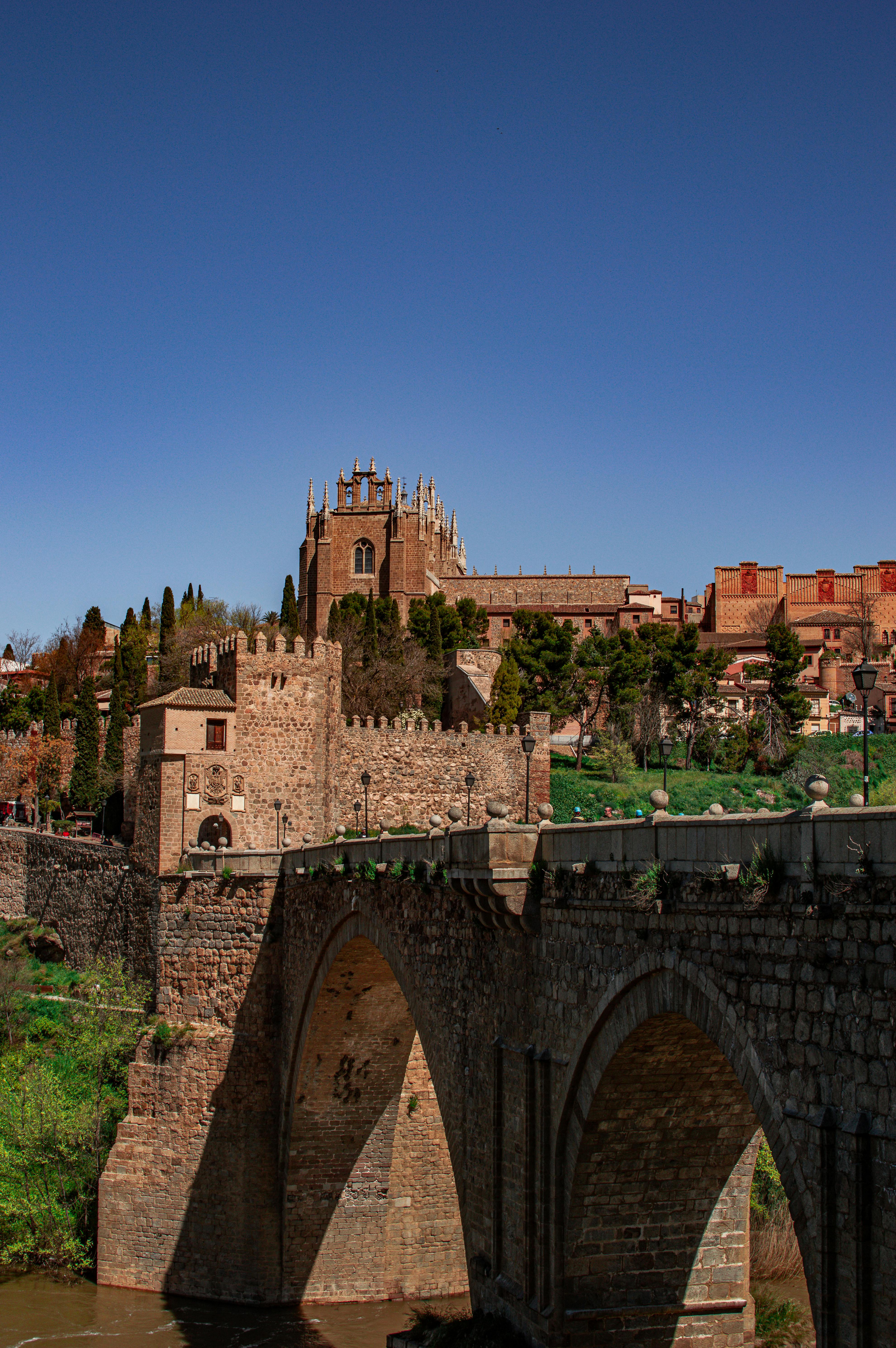 Scenic view of a medieval bridge with a historic cathedral in Toledo, Spain under a clear blue sky.
